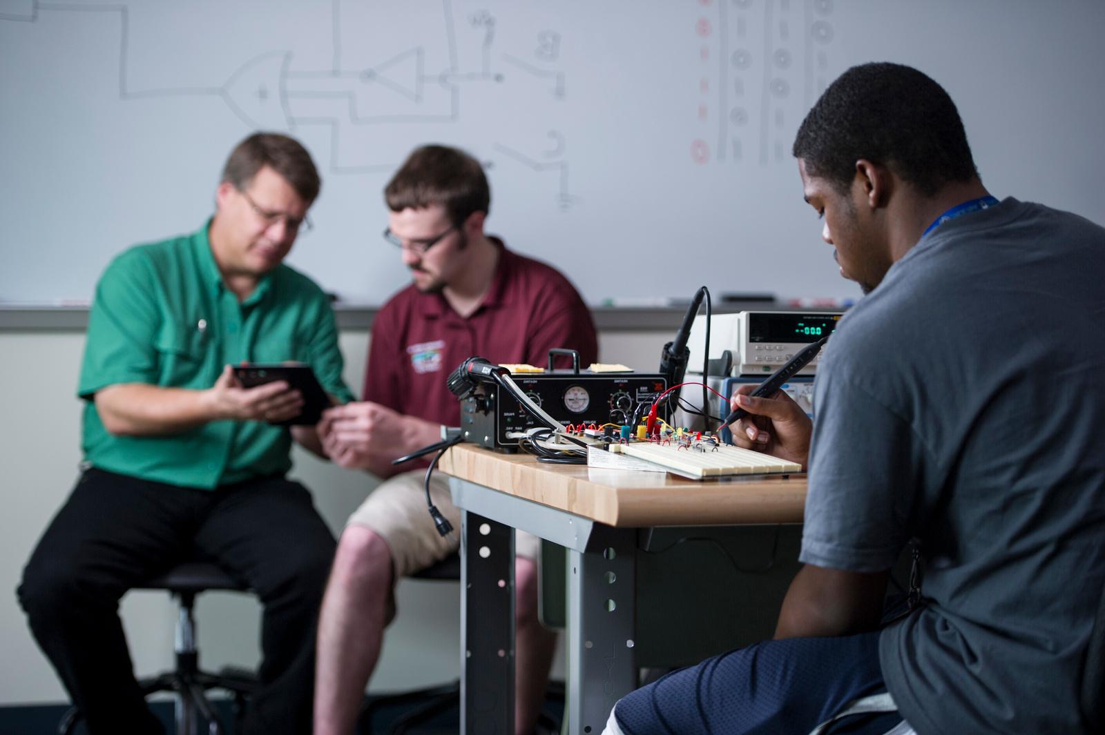 Teacher helps one student while another student uses a tool.